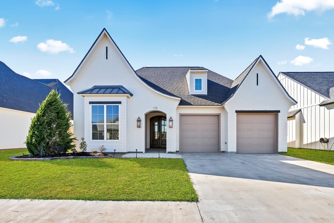 A single-story house with a pointed roof and beige exterior features two auto draft garage doors, a small front porch, and a neatly landscaped lawn. The sky is clear with a few clouds.