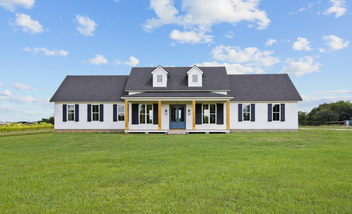 A single-story house with a dark roof and light-colored siding stands in the center of a grassy field, reminiscent of an auto draft illustration. The home boasts a symmetrical design with two dormer windows, a front porch, and a clear blue sky overhead.
