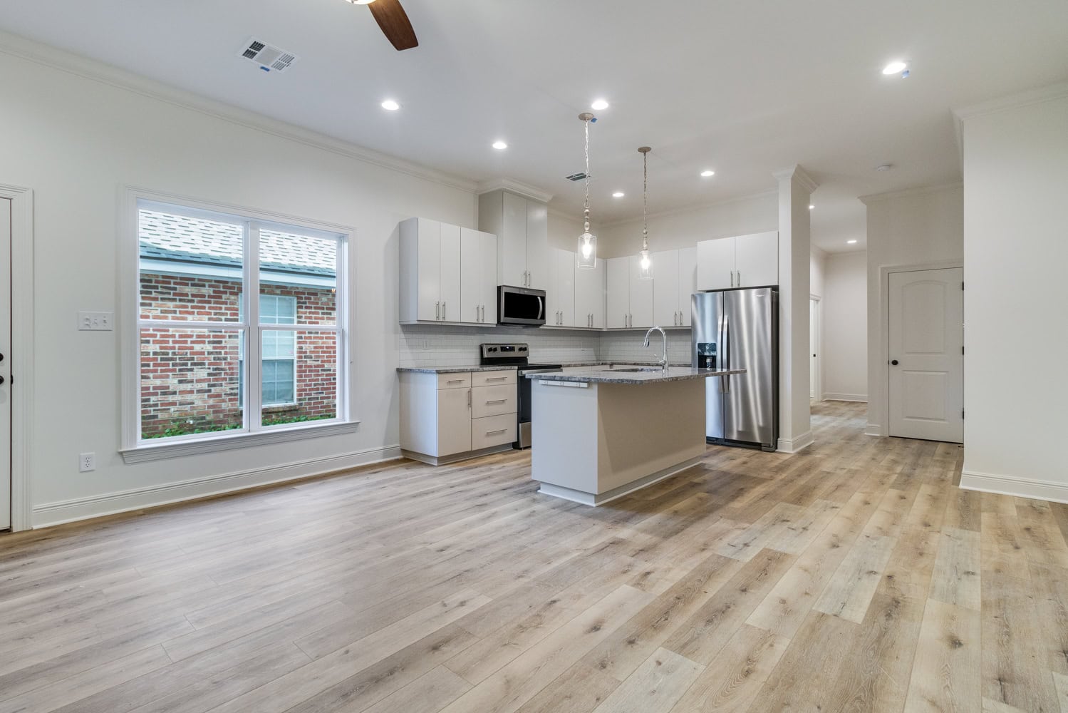 Modern kitchen and dining area with light wood floors, white cabinets, stainless steel appliances, a kitchen island, pendant lights, and a large window overlooking a brick wall outside. Room is well-lit and unfurnished.