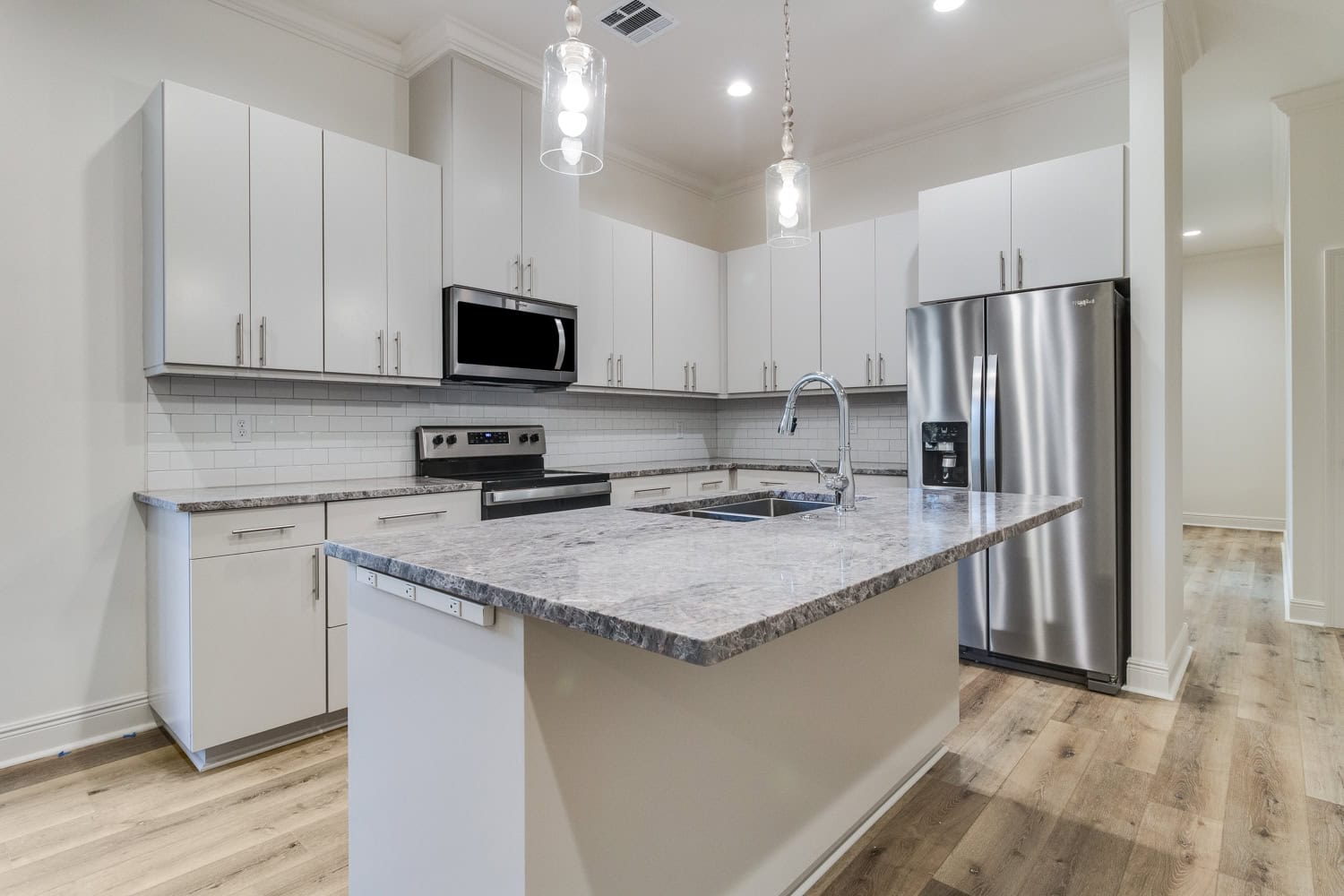 Modern kitchen with white cabinets, stainless steel appliances including a refrigerator and oven, an island with a sink and gray countertop, pendant lighting, light wood flooring, and a white subway tile backsplash.