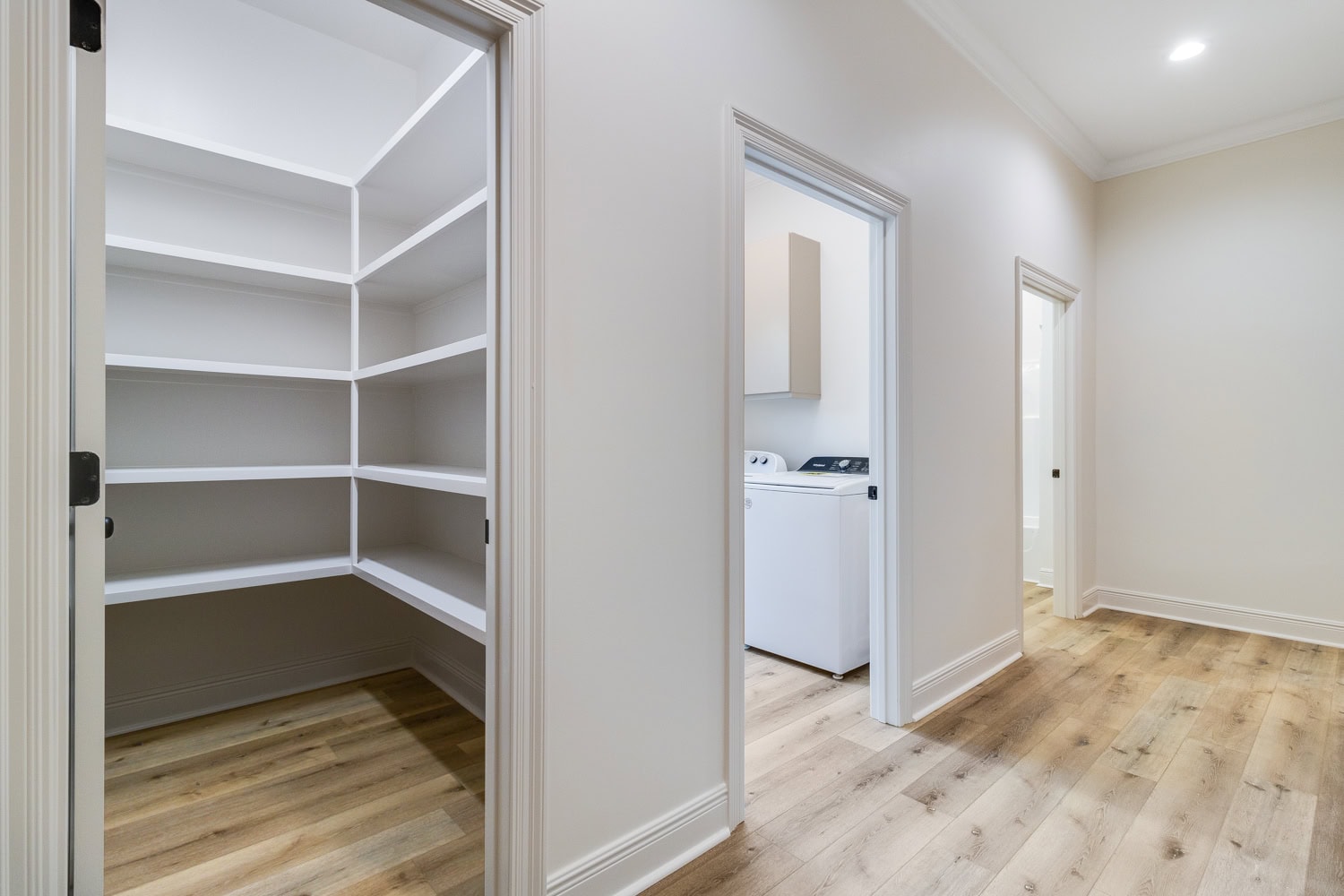 A hallway with light wood flooring features three doorways; one leads to a walk-in pantry with white shelves, another to a laundry room with a washer and dryer, and the third to a well-lit room.