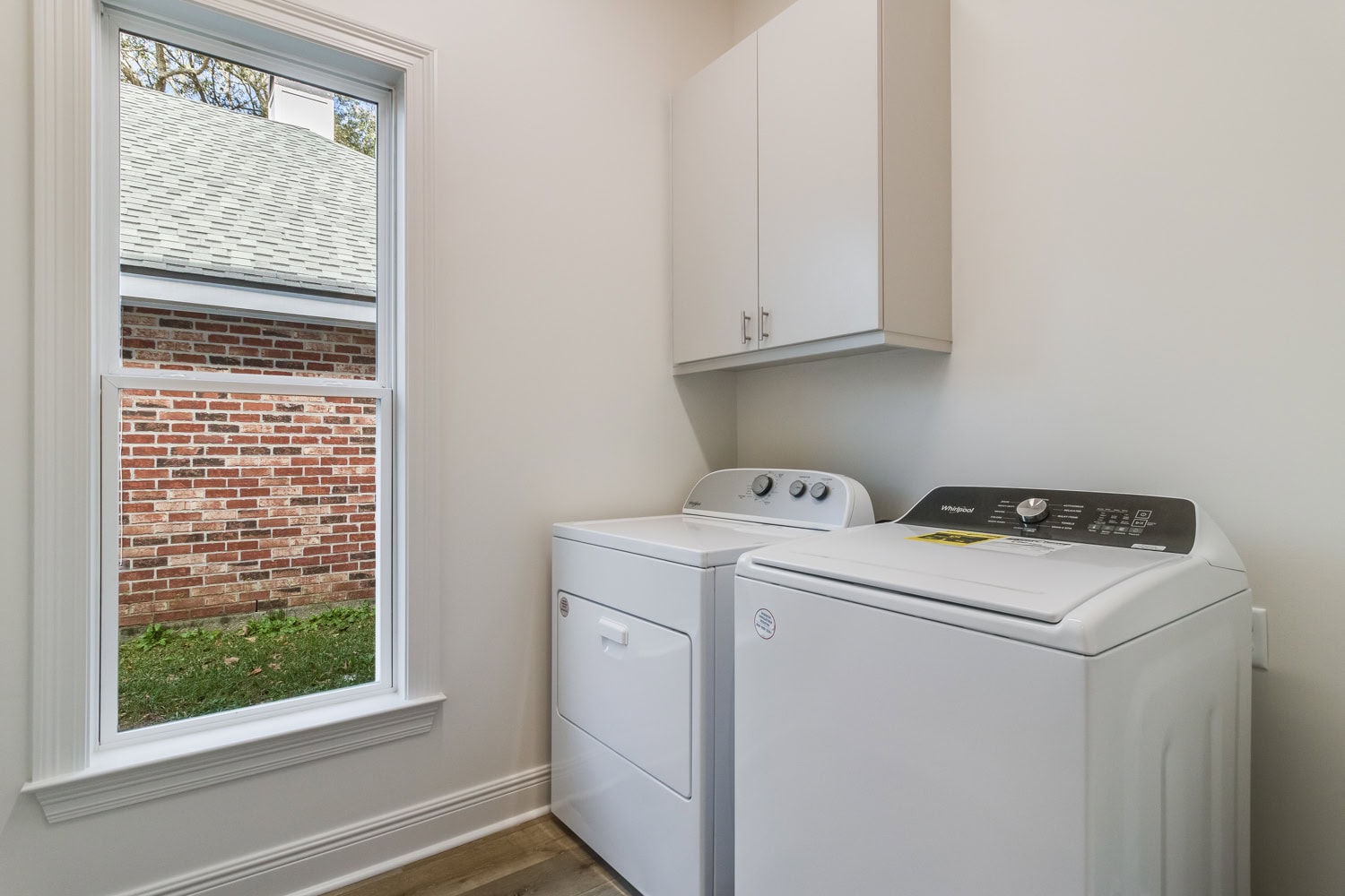A laundry room with a window, a white dryer and washer side by side, and a white cabinet mounted above them. The window shows an outside view of a brick wall and some grass.