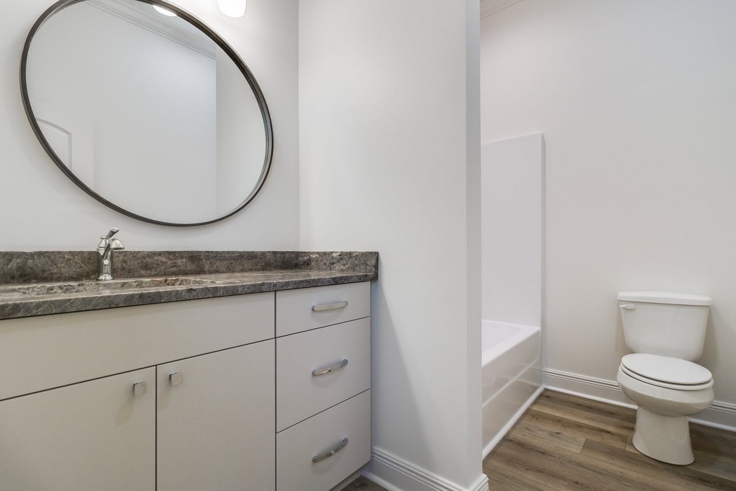 A bathroom with a marble countertop and sink, a large round mirror, white cabinets with silver handles, a toilet, and a bathtub with a white surround. The floor has wood-style flooring.