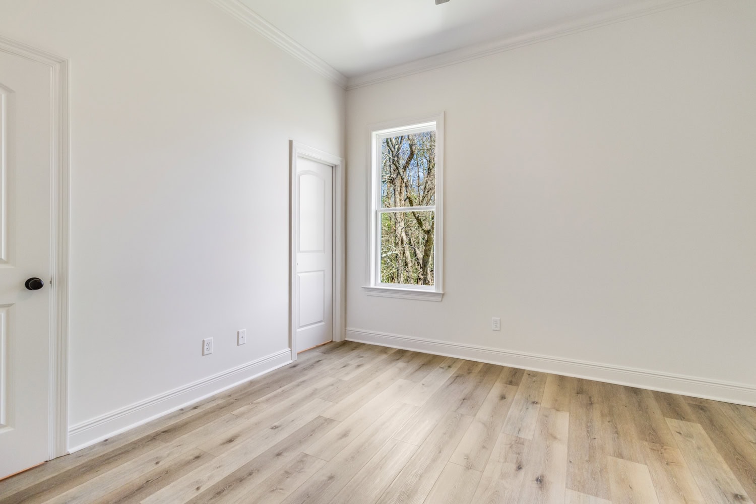 An empty room with light-colored walls, light wood flooring, a single window showing trees outside, and two white doors. The room has white trim and baseboards.
