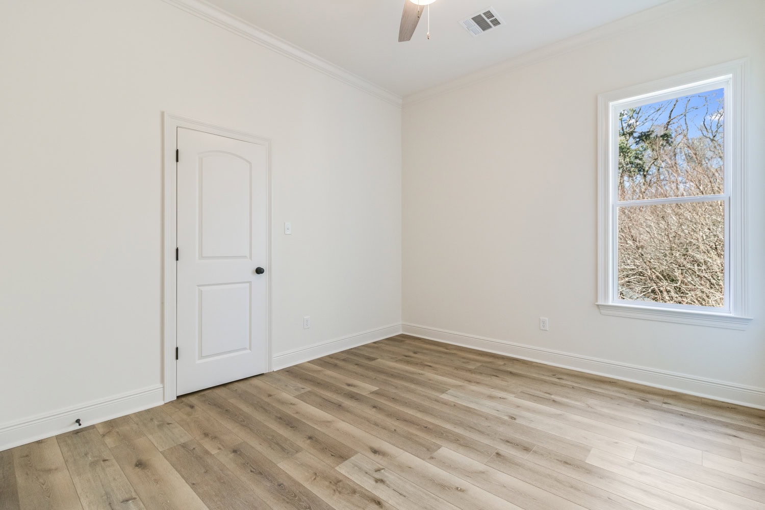 Empty room with light-colored walls, a white door, wood flooring, and a single window showing trees outside. Ceiling has a fan and air vent. The room is well-lit with natural light.