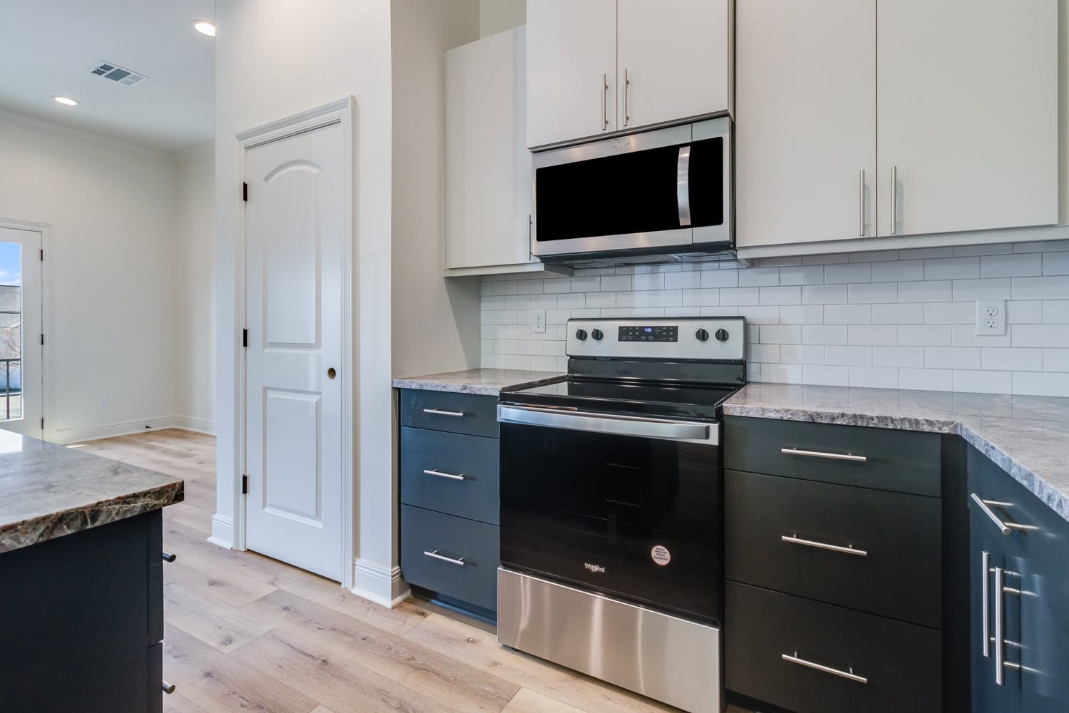 Modern kitchen with black and white cabinets, stainless steel oven and microwave, white subway tile backsplash, and light wood flooring. There is natural light coming from a door on the left.