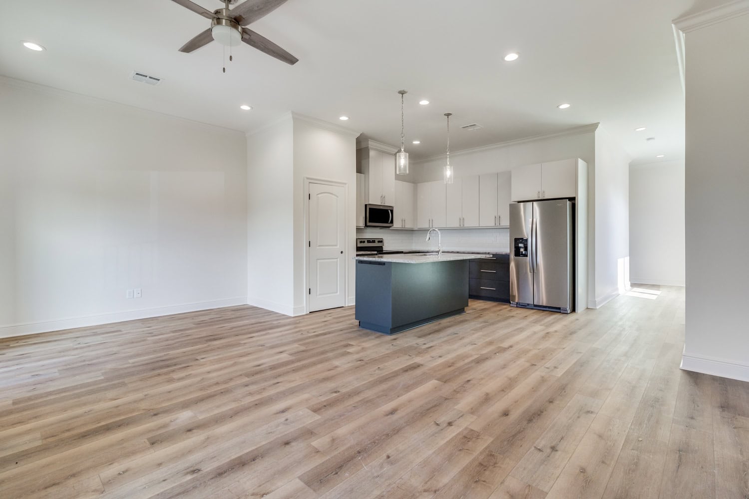 Modern, unfurnished open-concept kitchen and living area with wood flooring, white walls, ceiling fan, stainless steel appliances, pendant lights, and an island with dark cabinetry.