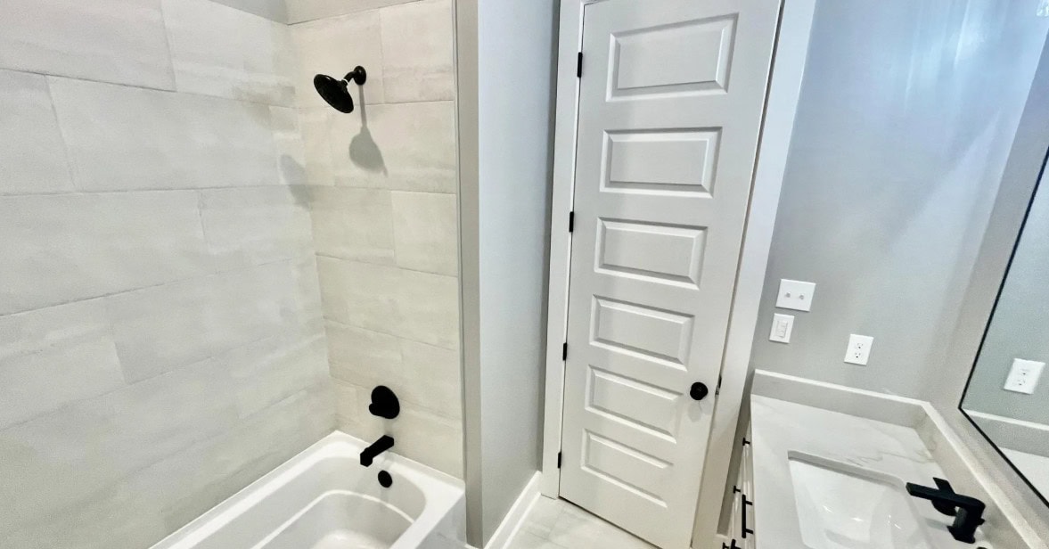 A bathroom with a white bathtub and shower on the left, a white paneled door in the center, and a sink with a rectangular mirror and black fixtures on the right. The walls and floor are light-colored.