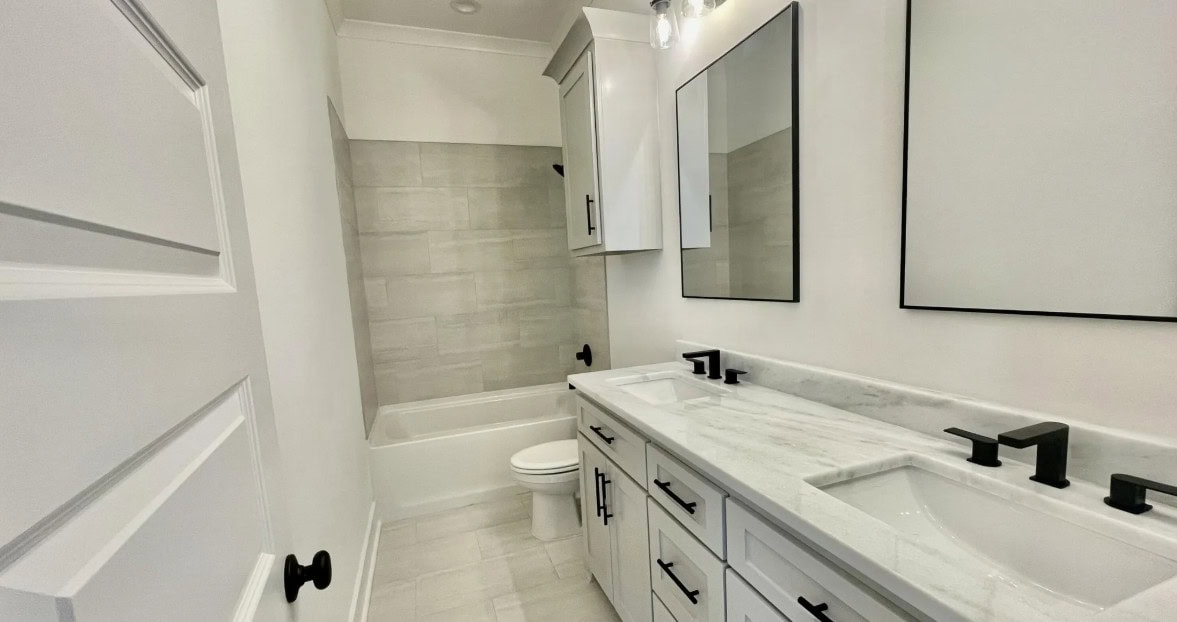 A modern bathroom with white walls, double sinks with black fixtures, a large mirror, and a bathtub with light gray tile. There are white cabinets above the toilet and under the sinks.