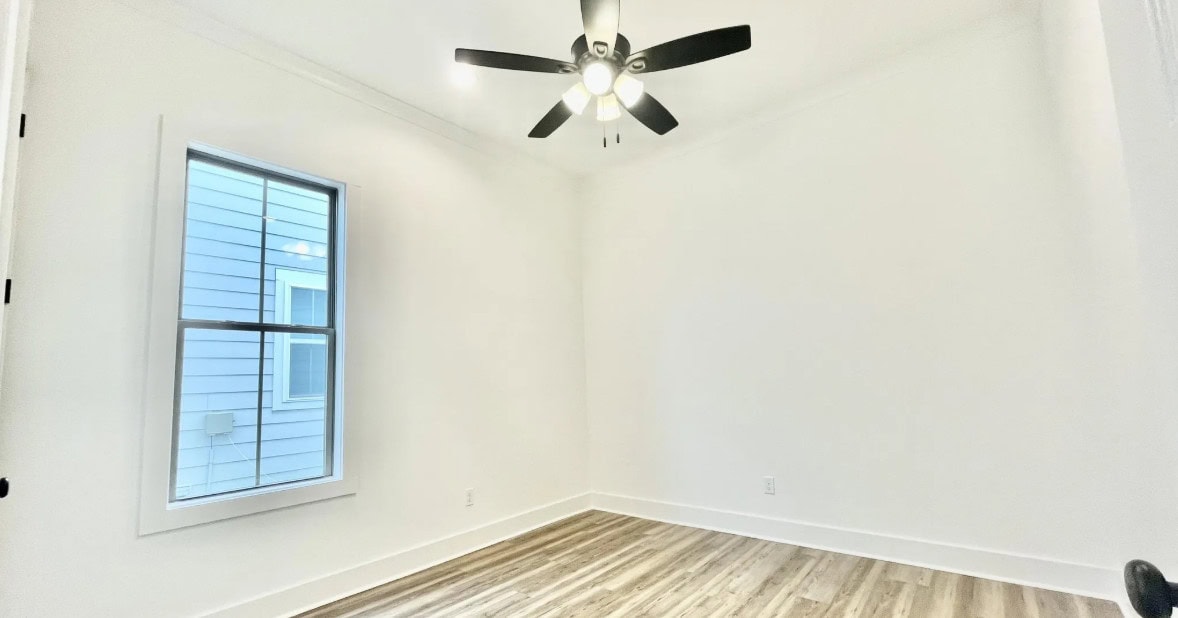 A small, empty room with white walls, a single window with a grid pattern, light wood flooring, and a black ceiling fan with lights. The room has minimal trim and is brightly lit.