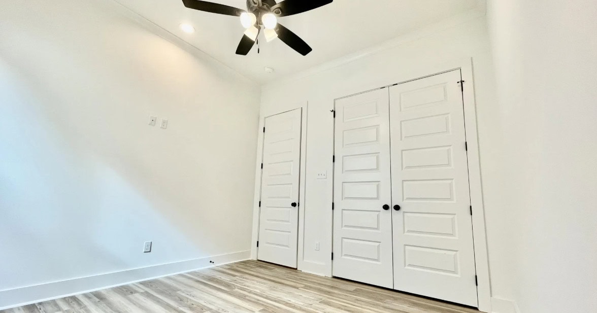 An empty room with white walls, a ceiling fan with lights, wood-patterned flooring, and two sets of double doors with black knobs. The room is brightly lit by ceiling lights.