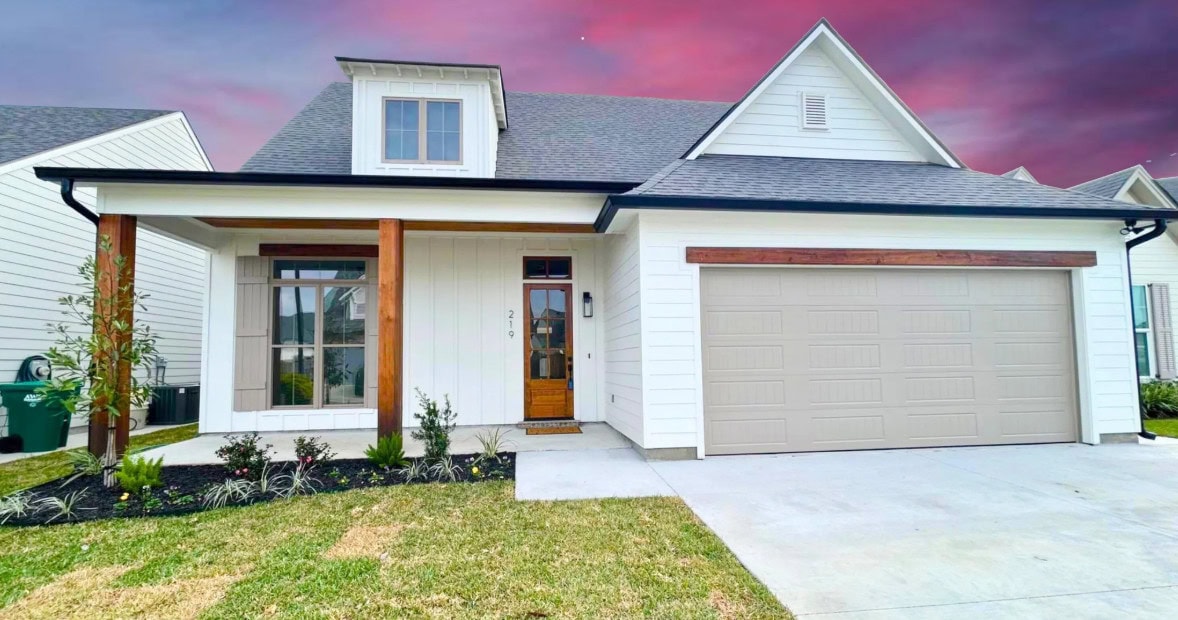 A modern white house with wooden accents, a covered front porch, two large front windows, a light brown garage door, and a well-maintained lawn, set against a purple and pink sunset sky.