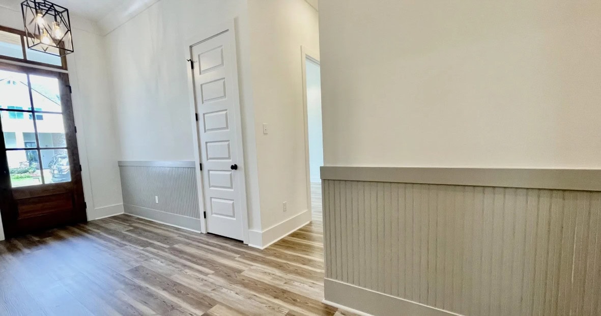 A bright hallway with wood flooring and white walls, featuring wainscoting and a white paneled door. Natural light enters from a window near a wooden front door.
