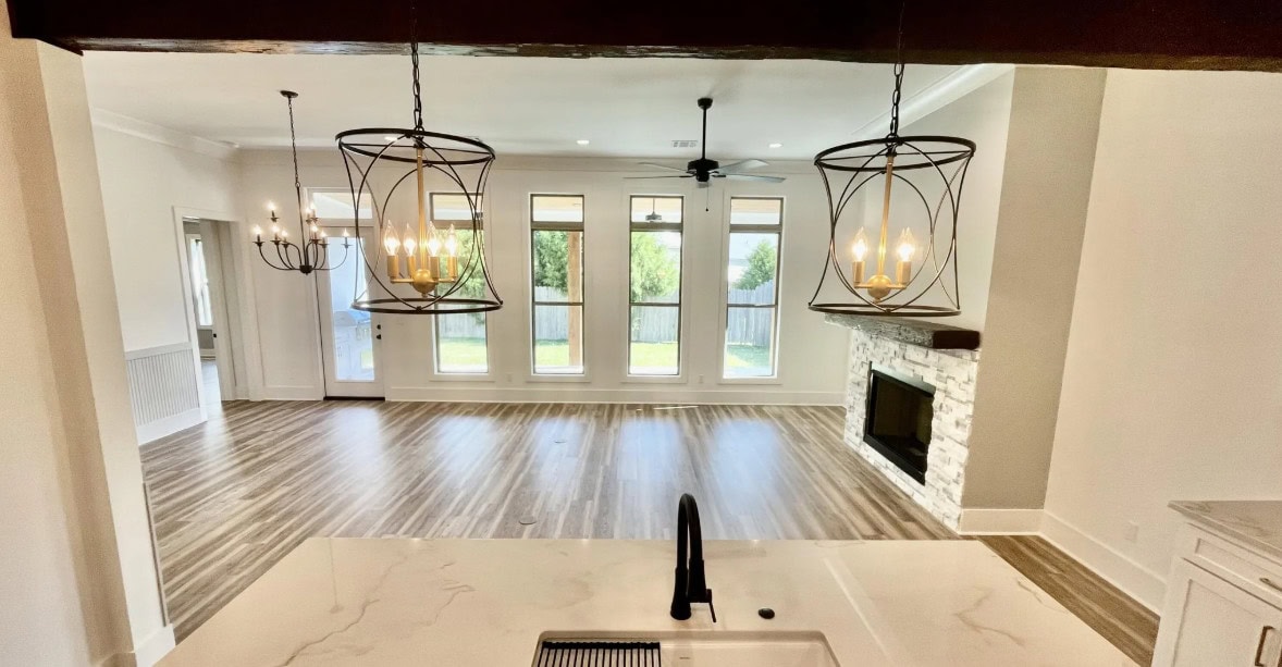 A view from a kitchen sink looking into an open living area with wood flooring, large windows, a stone fireplace, and two modern pendant lights hanging from the ceiling. A chandelier is visible in the adjacent dining space.
