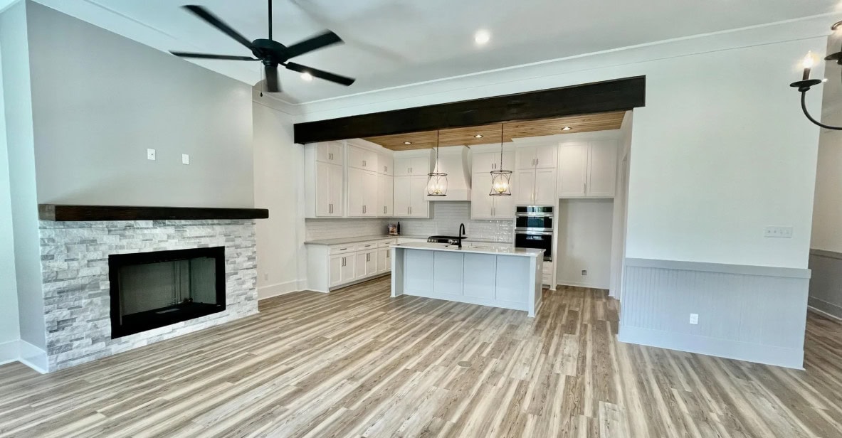 A modern kitchen with white cabinets, an island, pendant lights, and built-in appliances, adjacent to a living area with a stone fireplace, ceiling fan, and wood-patterned flooring.