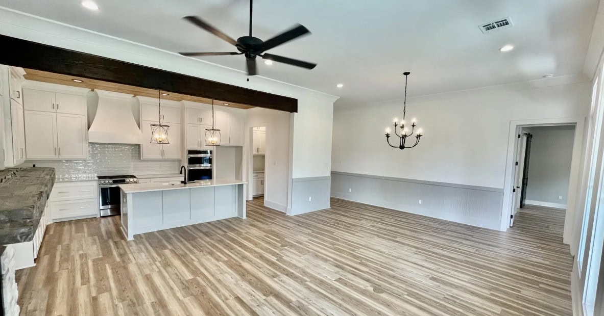 A spacious, empty open-concept kitchen and living area with light wood flooring, white cabinets, a large kitchen island, modern pendant and chandelier lighting, and a ceiling fan.