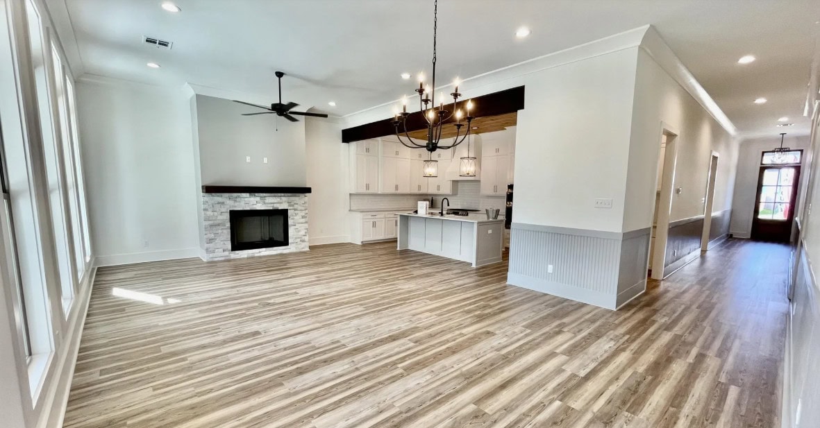 Open-concept living and kitchen area with wood-style flooring, white walls, a brick fireplace, large chandelier, ceiling fan, and an island; hallway leads to the front door with glass panels.