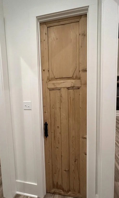 A narrow, unfinished wooden door with a rectangular panel design, set in a white door frame, with a black handle on the left side and a light switch on the adjacent wall.