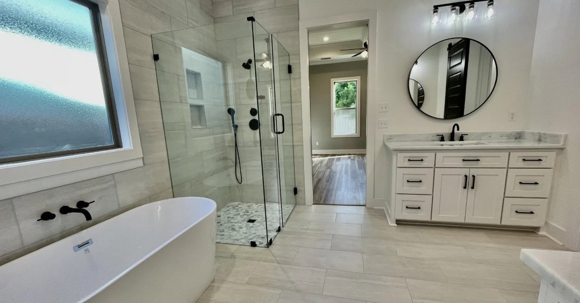 Modern bathroom with a glass-enclosed shower, a freestanding bathtub, white cabinets with a marble countertop, a round mirror, and a window letting in natural light. Light-colored tile flooring throughout.