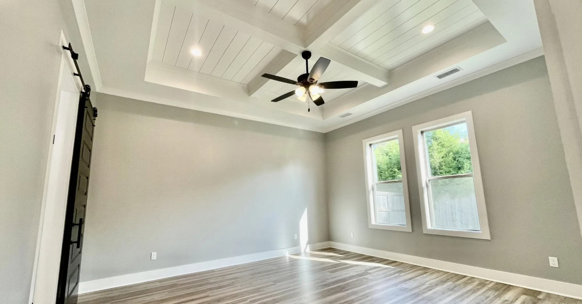 A bright, empty room with light gray walls, wood flooring, two large windows, a ceiling fan with lights, and a coffered white ceiling with recessed lighting. A black sliding barn door is on the left wall.