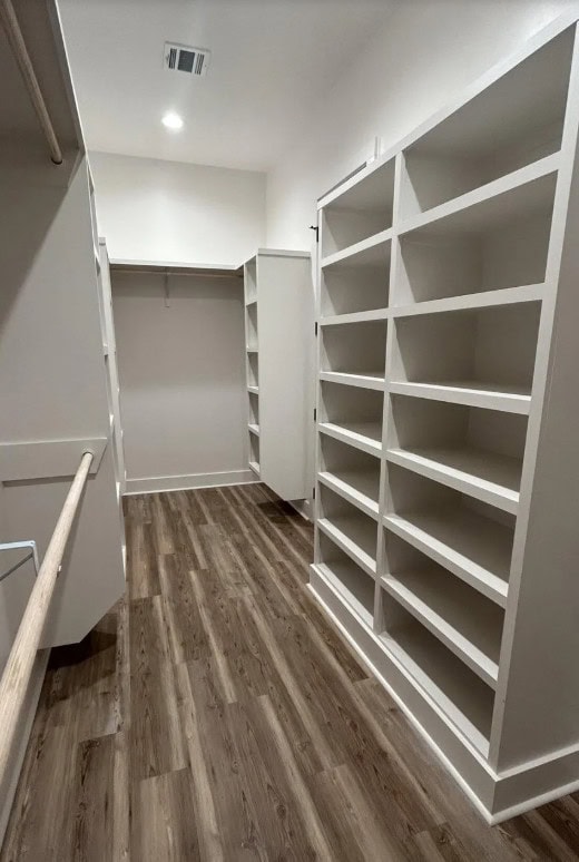 A walk-in closet with empty built-in shelves and hanging rods on both sides, white walls, and wood-patterned flooring under recessed ceiling lighting.