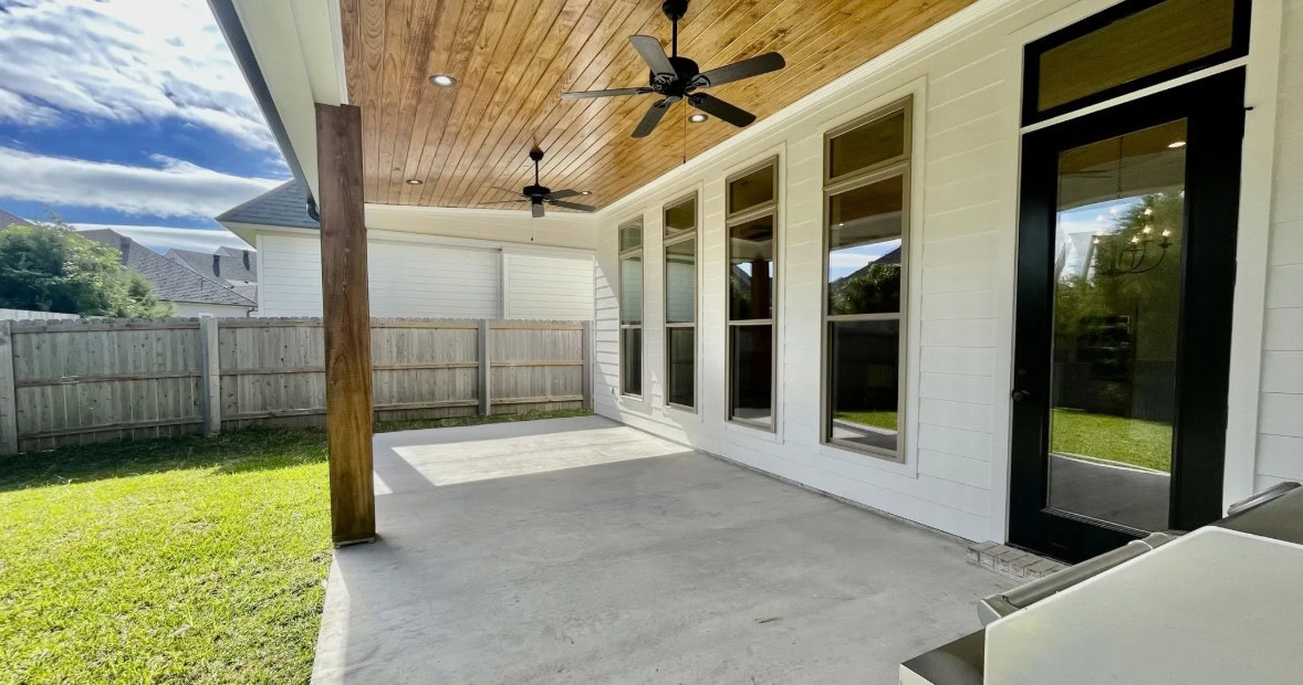 A covered backyard patio with a wood-paneled ceiling, two ceiling fans, large windows, a black-framed glass door, and a concrete floor. A wooden fence surrounds a grassy yard under a partly cloudy sky.