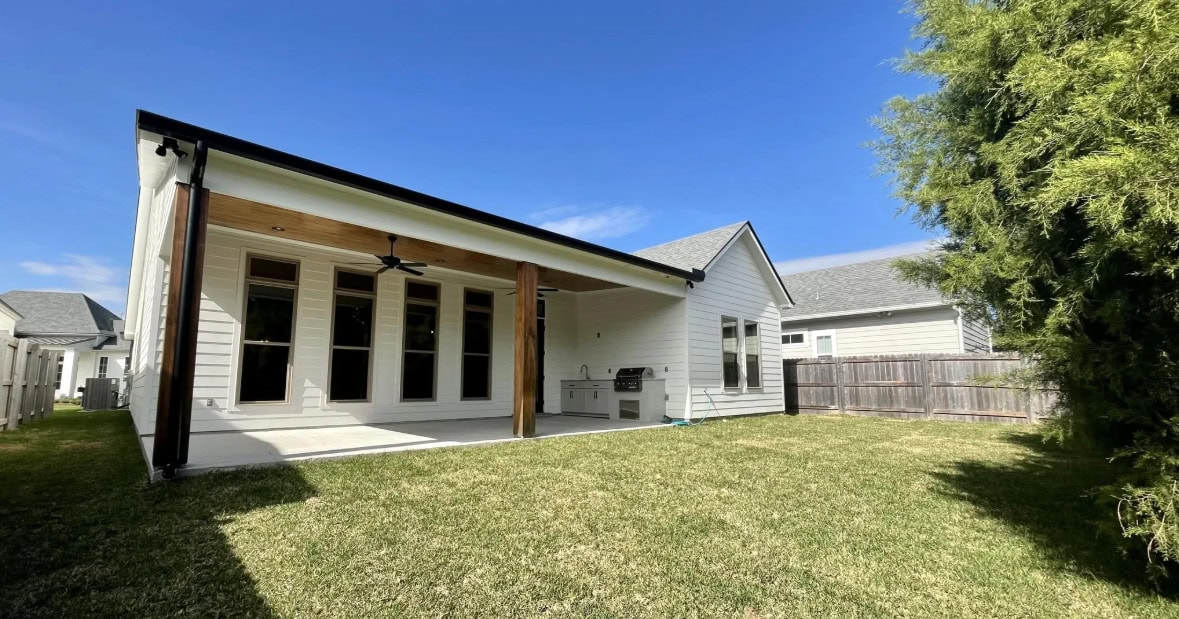A modern white house with a covered patio, ceiling fan, outdoor kitchen, and large windows. The backyard features a green lawn, wooden fence, and a tall tree on the right side under a clear blue sky.