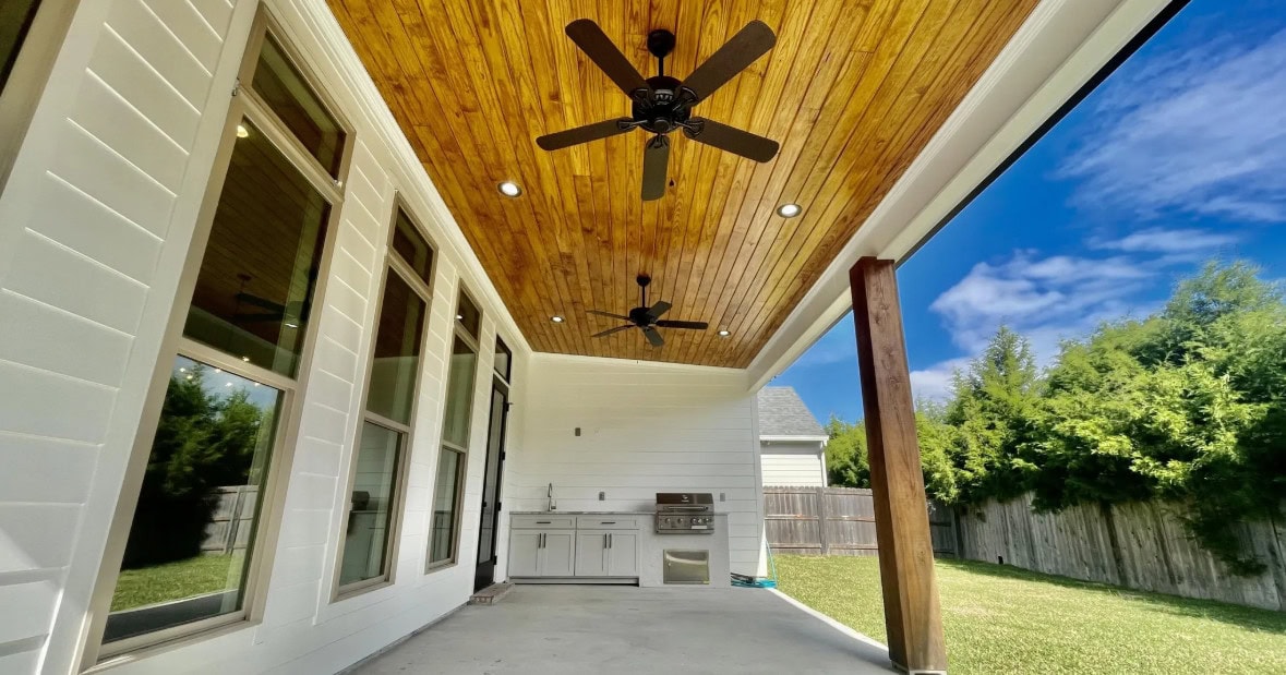 A covered outdoor patio with a wooden ceiling, two ceiling fans, recessed lights, a built-in grill, white cabinetry, and large windows. The patio overlooks a green lawn fenced in by wooden panels.