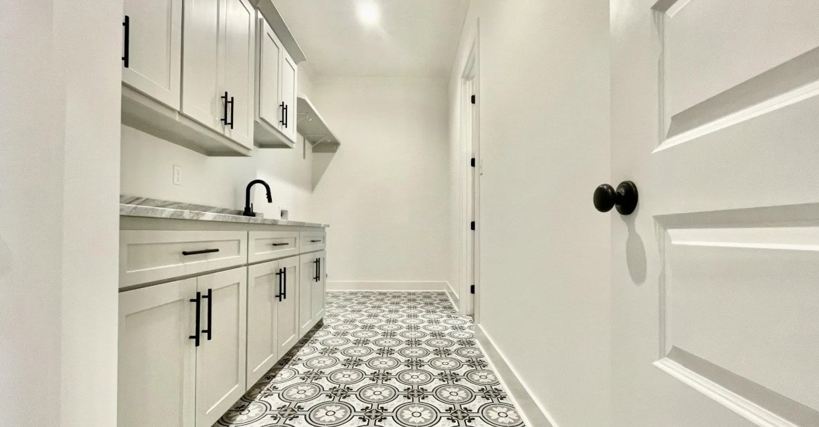 A narrow, white utility room with patterned tile flooring, light gray cabinets, black hardware, a countertop with a black faucet, and several closed doors along the walls.