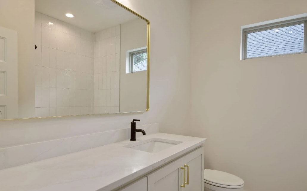 A modern bathroom with a white countertop, rectangular sink, dark faucet, large wall mirror with a gold frame, white cabinets, a toilet, and a small window letting in natural light.