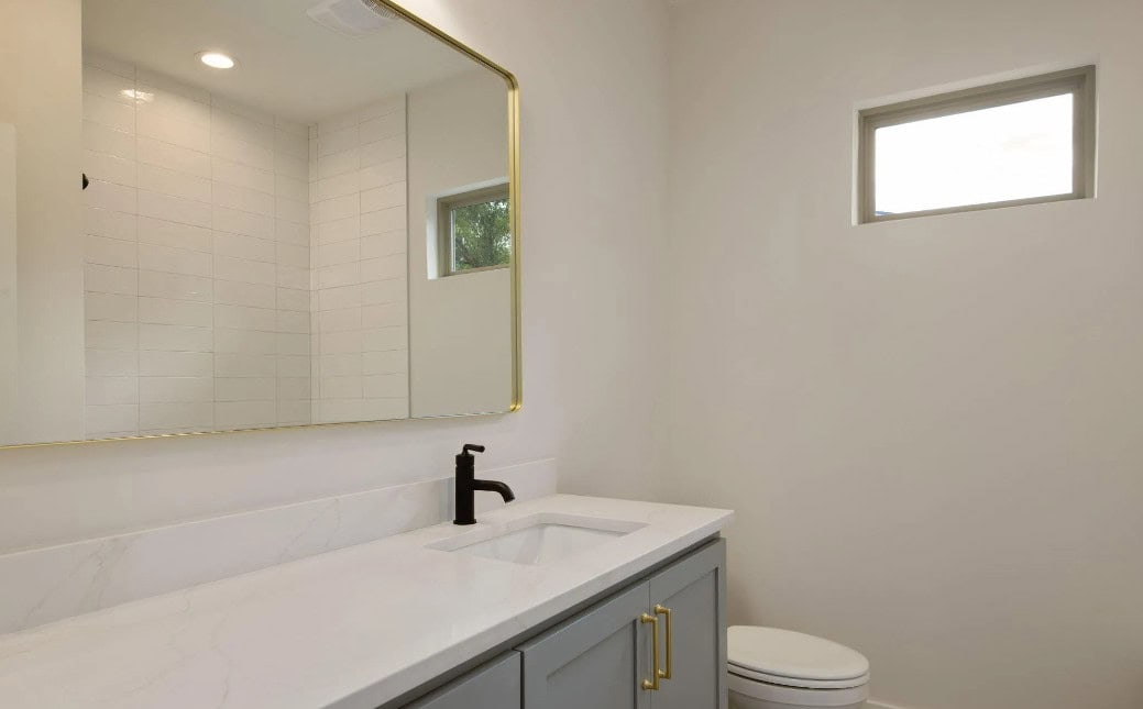 A modern bathroom with a large rectangular mirror above a white countertop, a black faucet, grey cabinets with gold handles, a toilet, white tiled shower, and a small window on the right wall.