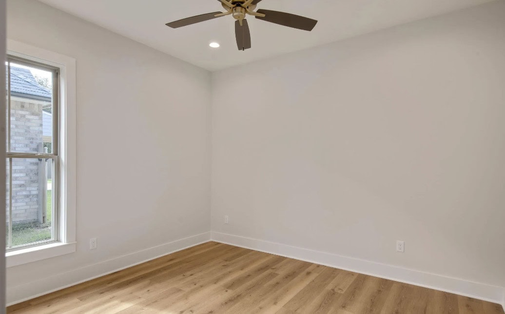An empty room with light-colored walls, a ceiling fan with five blades, wood-look flooring, a single window on the left letting in natural light, and white baseboards.