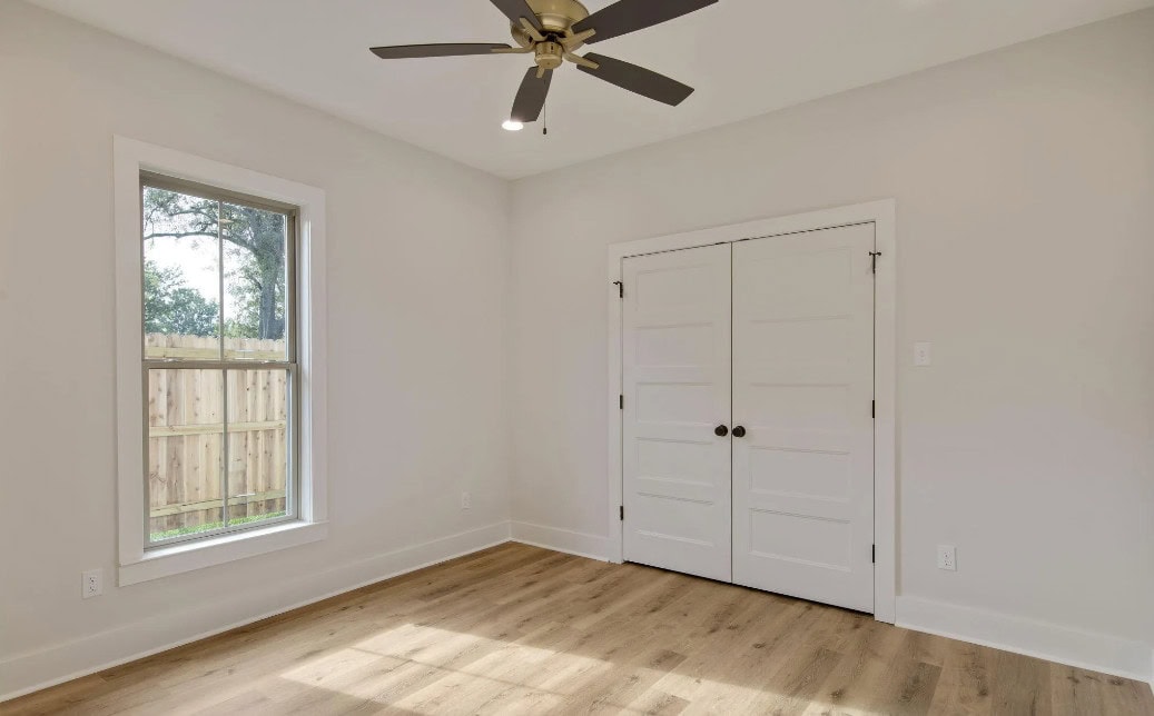 A bright, empty room with light-colored walls and wood flooring, a window on the left, double white doors on the right wall, and a ceiling fan with a light fixture above.