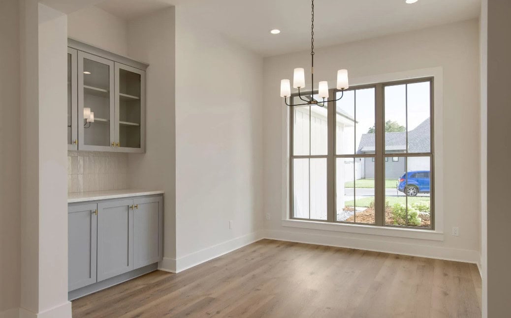 A bright, empty room with light wood floors, a large window with sheer curtains, a modern chandelier, and built-in gray cabinets with glass doors on the left wall. A blue car is visible outside the window.