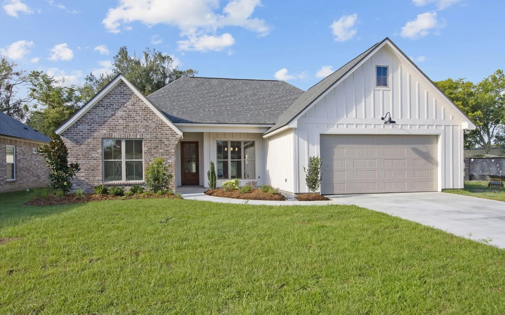A single-story suburban house with brick and white siding, a front porch, gray shingle roof, attached two-car garage, manicured lawn, and shrub landscaping under a partly cloudy sky.