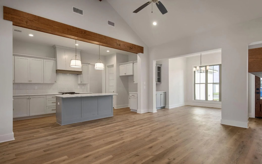 A modern kitchen with light gray cabinets, white backsplash, a center island with pendant lights, wood beam accents, wood flooring, and an adjacent open dining area with large windows.