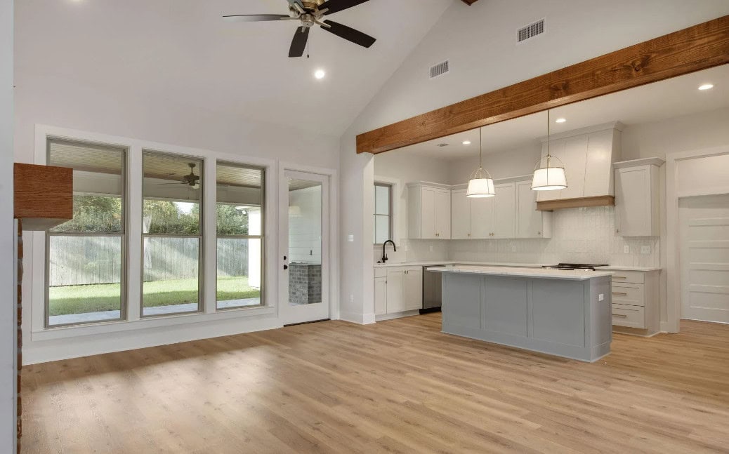 A spacious, empty kitchen and living area with light wood floors, large windows, vaulted ceiling with exposed wooden beam, white cabinetry, pendant lights over a gray island, and a ceiling fan.