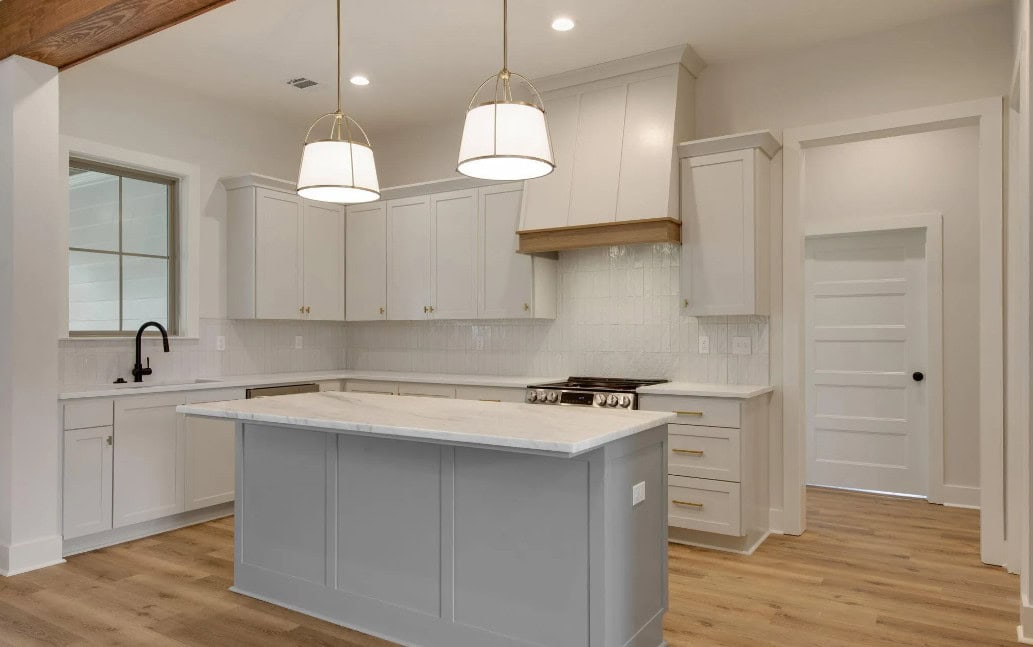 A modern kitchen with light gray cabinets, a matching center island, white countertops, two pendant lights, a built-in stove with a range hood, wood flooring, and a white door in the background.