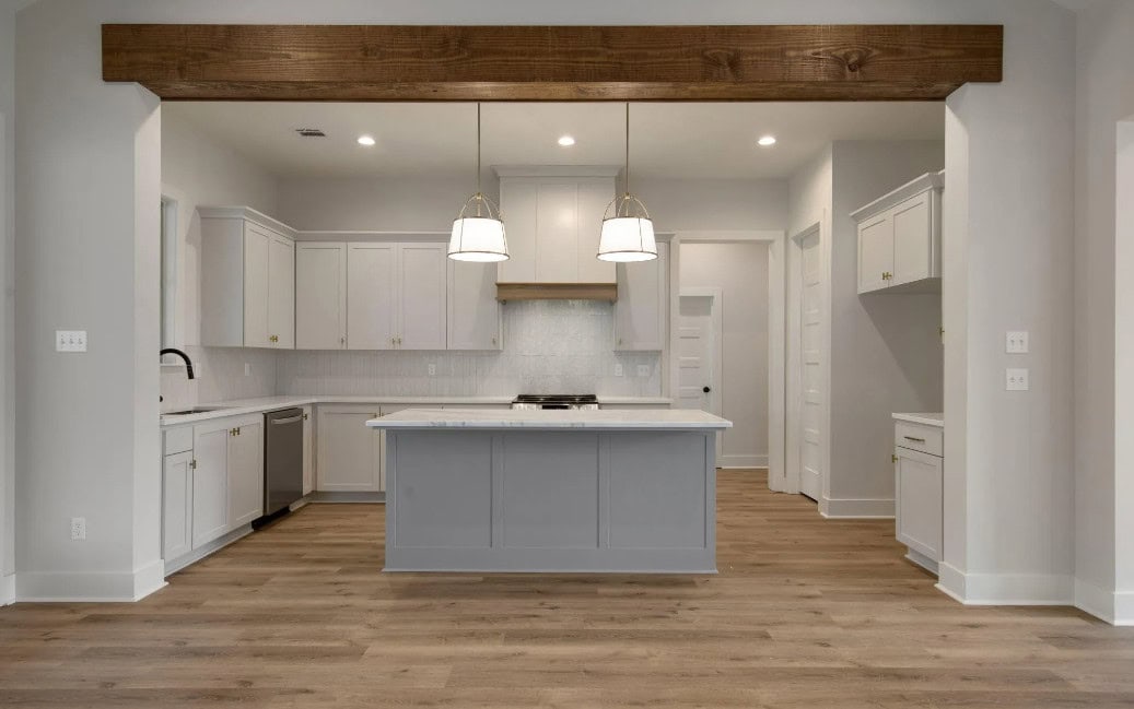 Modern kitchen with light gray cabinets, a central island, stainless steel appliances, white backsplash, wood floors, two pendant lights, and a large wooden beam across the ceiling.