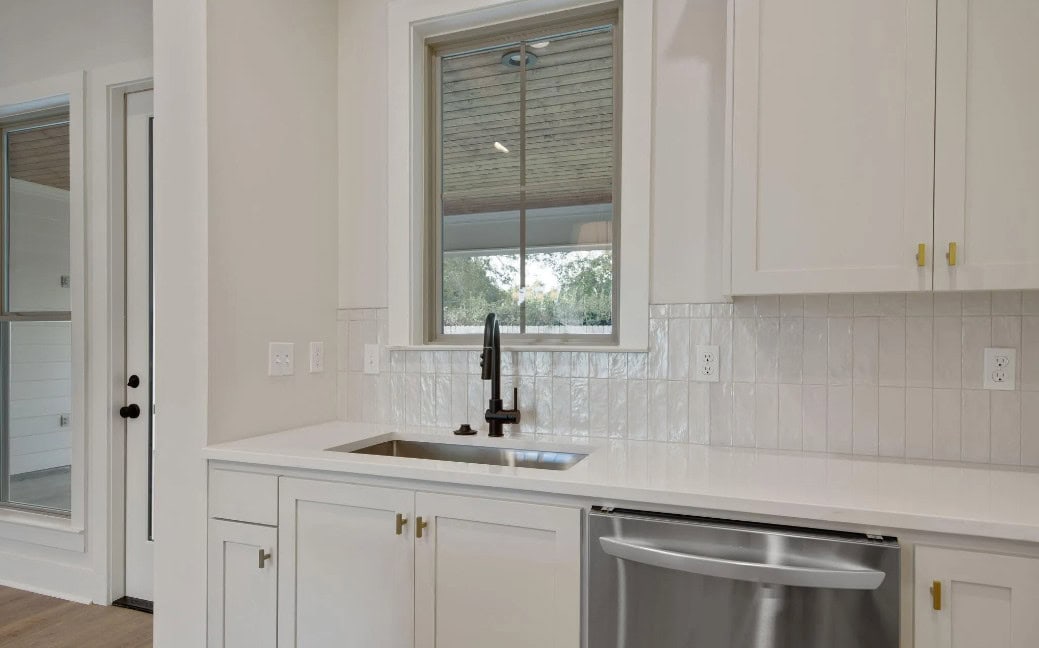 A modern kitchen features white cabinets, a stainless steel dishwasher, a black faucet, and a rectangular sink beneath a window. The backsplash is made of white tiles, and there are multiple electrical outlets on the wall.