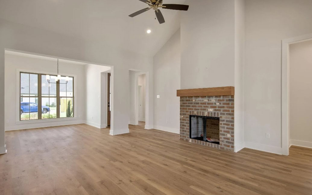 A spacious, empty living room with light wood flooring, a brick fireplace with a wooden mantle, large window, ceiling fan, and adjoining dining area under a modern chandelier. Walls are painted off-white.