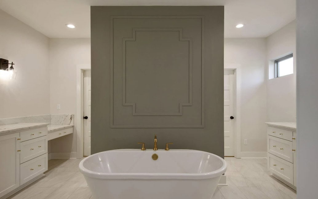 A white freestanding bathtub with gold fixtures is positioned in front of a gray accent wall. The bathroom features white cabinets, marble countertops, light tile flooring, and recessed ceiling lights.