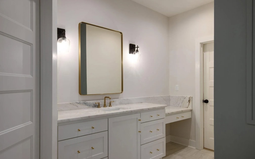 A bathroom vanity with white cabinets, gold handles, a marble countertop, a large rectangular mirror with a gold frame, two wall-mounted lights, and a door to the right.