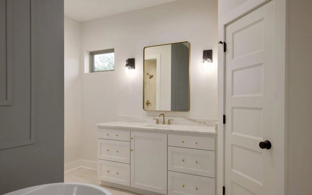 A modern bathroom with a white vanity, gold hardware, and a marble countertop. A rectangular mirror is above the sink, flanked by two wall-mounted lights. A door and a small window are also visible.