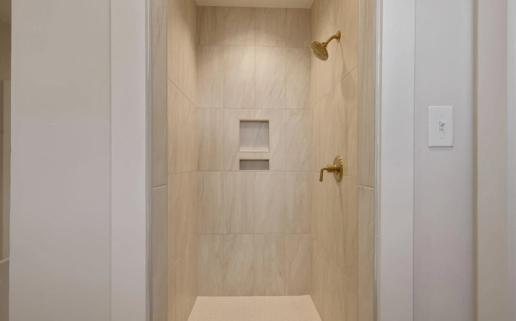 A beige tiled walk-in shower with two showerheads on the right wall, a built-in recessed shelf in the center, and a white wall switch visible on the right outside the shower.
