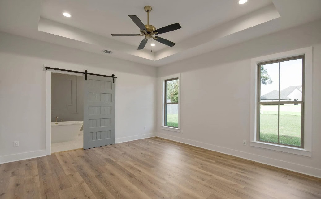 A bright, empty room with light wood flooring, two large windows, a tray ceiling with recessed lights, a black ceiling fan, and a sliding barn door leading to a bathroom with a bathtub.