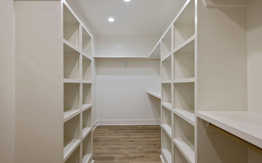 A walk-in closet with white built-in shelves on both sides, wooden rods for hanging clothes, and a light wood floor. The space is empty and well-lit with ceiling lights.