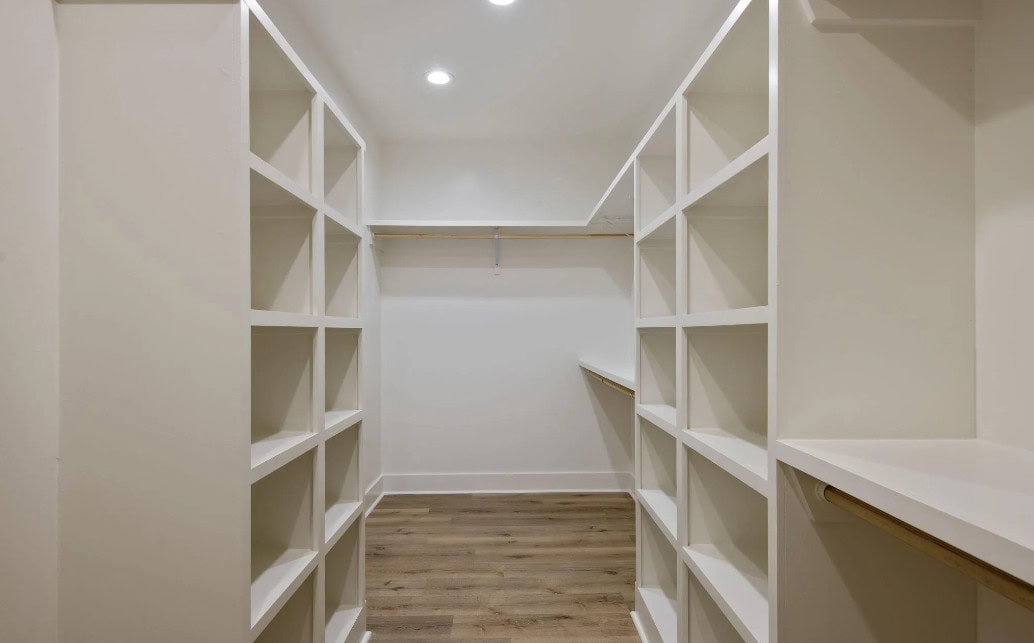 A walk-in closet with white built-in shelves on both sides, a wooden rod for hanging clothes, and wood flooring. The room is well-lit with recessed ceiling lights.