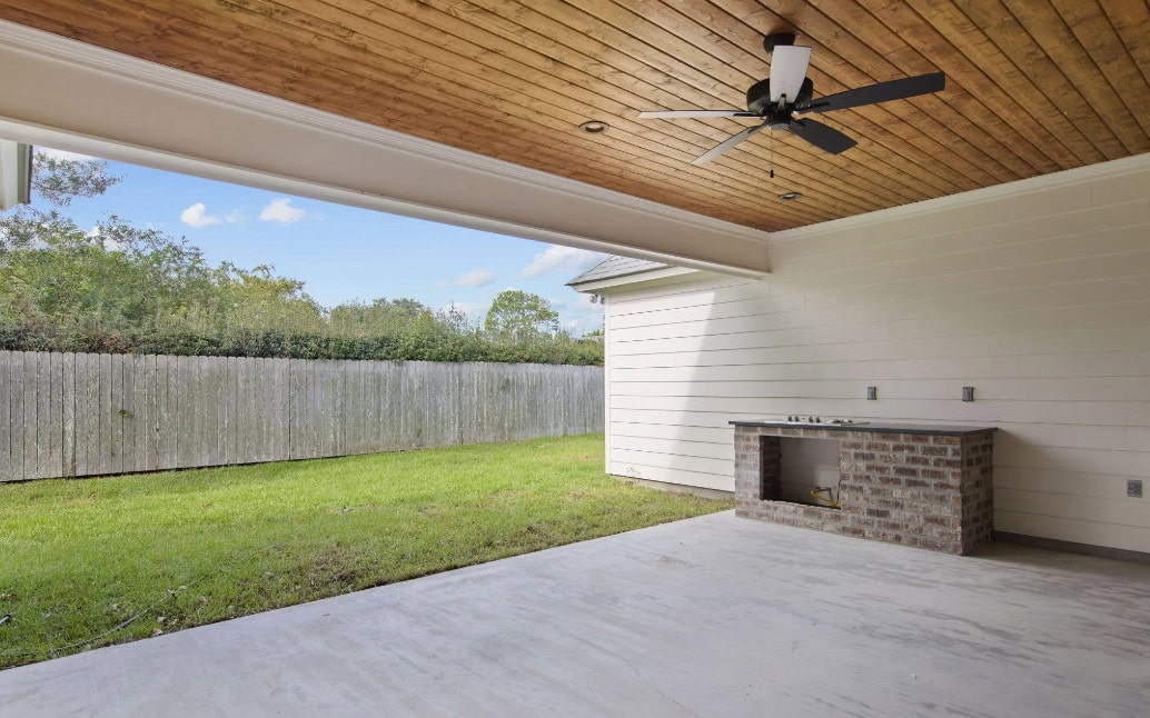 A covered patio with a wood-paneled ceiling, ceiling fan, concrete floor, brick outdoor kitchen area, and a fenced backyard with grass and trees. The sky is partly cloudy.