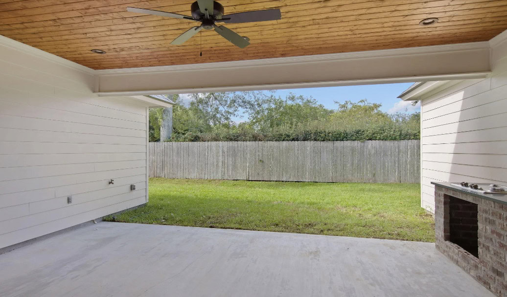 A covered patio with a wooden ceiling and ceiling fan overlooks a grassy backyard enclosed by a wooden privacy fence. There is a built-in brick feature on the right and wall outlets on the left.